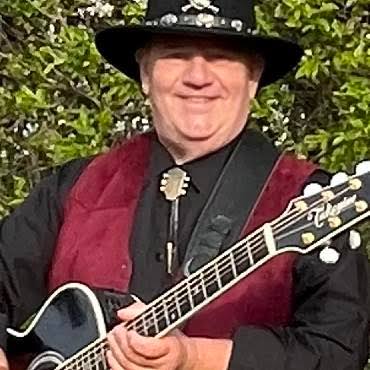 The Singing Cowboy in a black hat and red waistcoat playing an acoustic guitar